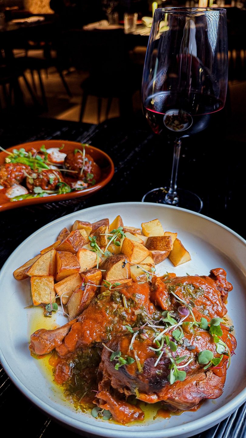Braised meat with roasted potatoes and a glass of red wine on a dark table.