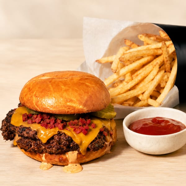 A double smash burger with cheese, bacon bits, pickles, fries, and ketchup on a wooden table.